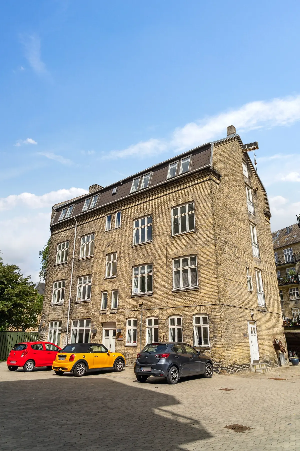 tre biler parkeret foran et stort lejlighedsbyggeri med røde mursten og hvide ruder