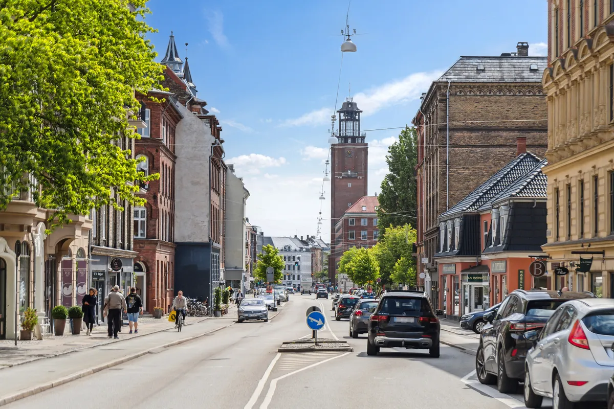 Mennesker går på gågaden i København og kører biler og cykler langs en gade med bygninger og træer og en klokketårn på