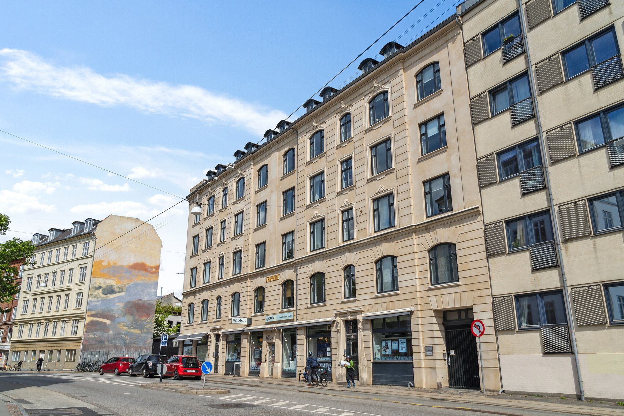 Gade med forretninger og biler parkeret langs med en stor graffitimur, der viser landskaber