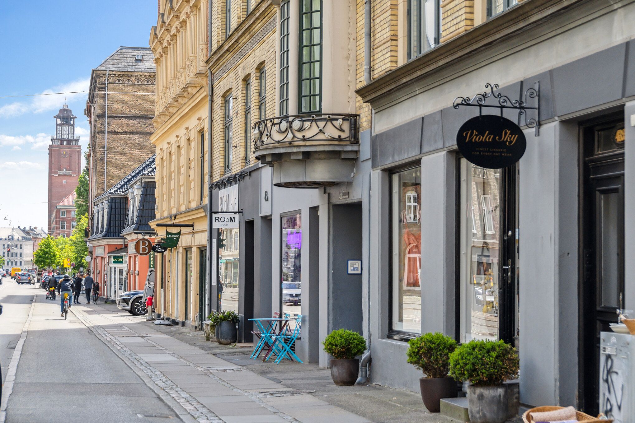 Lejemejl i centrum af København med smukke butikker, grønne planter og en turistbygning med klokketårn i baggrunden