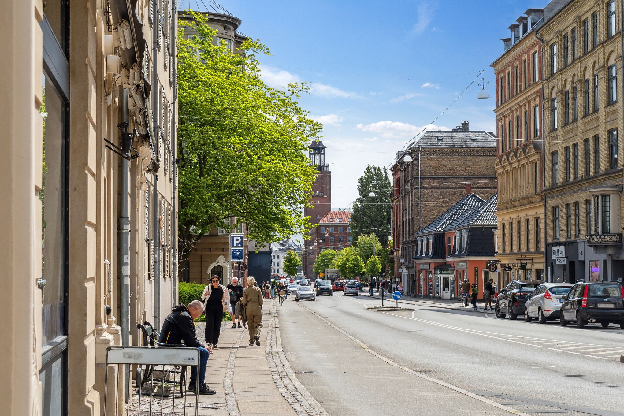 Flere mennesker går på gaden med skrånende sider og parkerede biler, mens en person sidder og læser på siden