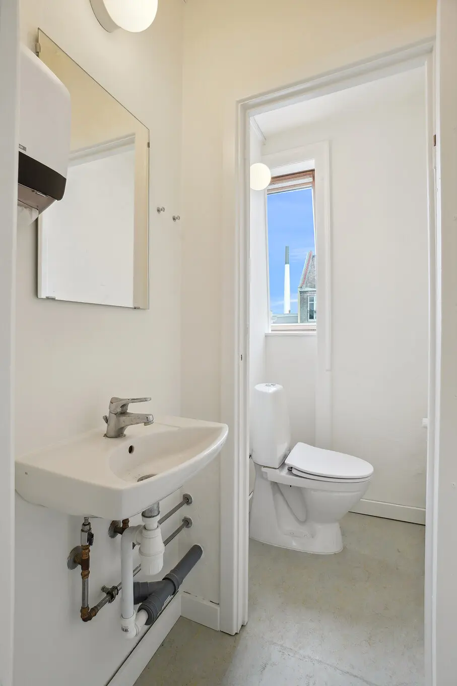 Et lille badeværelse med toilet, hvidt keramikbad og håndvask samt et spejl og en håndklædedispenser på væggen