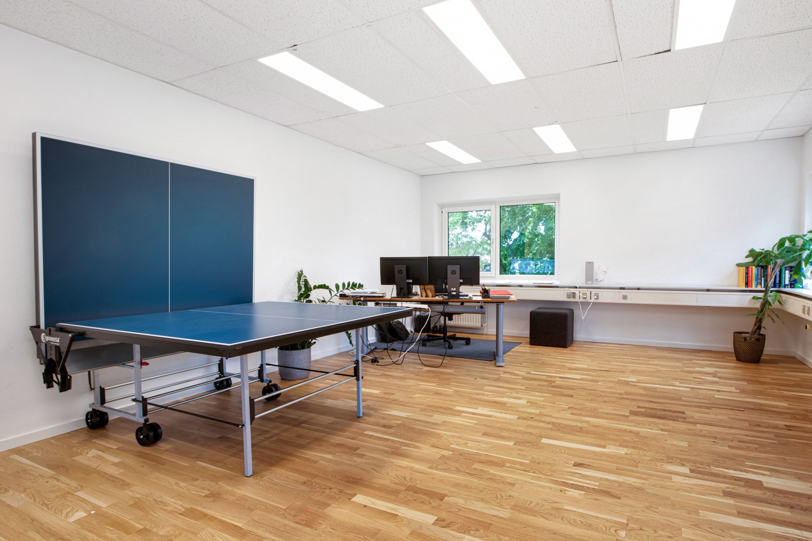 Kontor med bord, stole, computere, trægulv, en pingpongbord og plante i potte samt en vinduesvisning med grønne træer