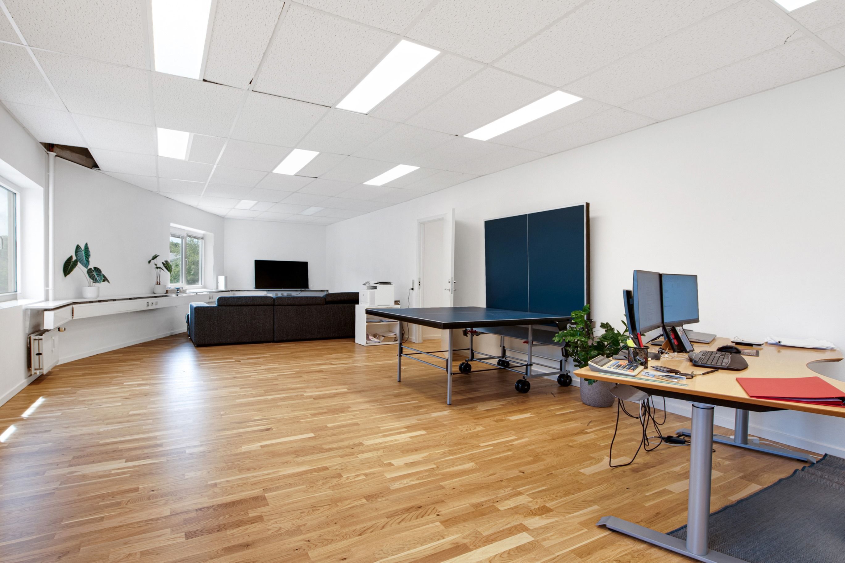 To skriveborde med computere og kalendere står i hjørnet af et kontorlokale med et pingpongbord og en sofa i baggrunden