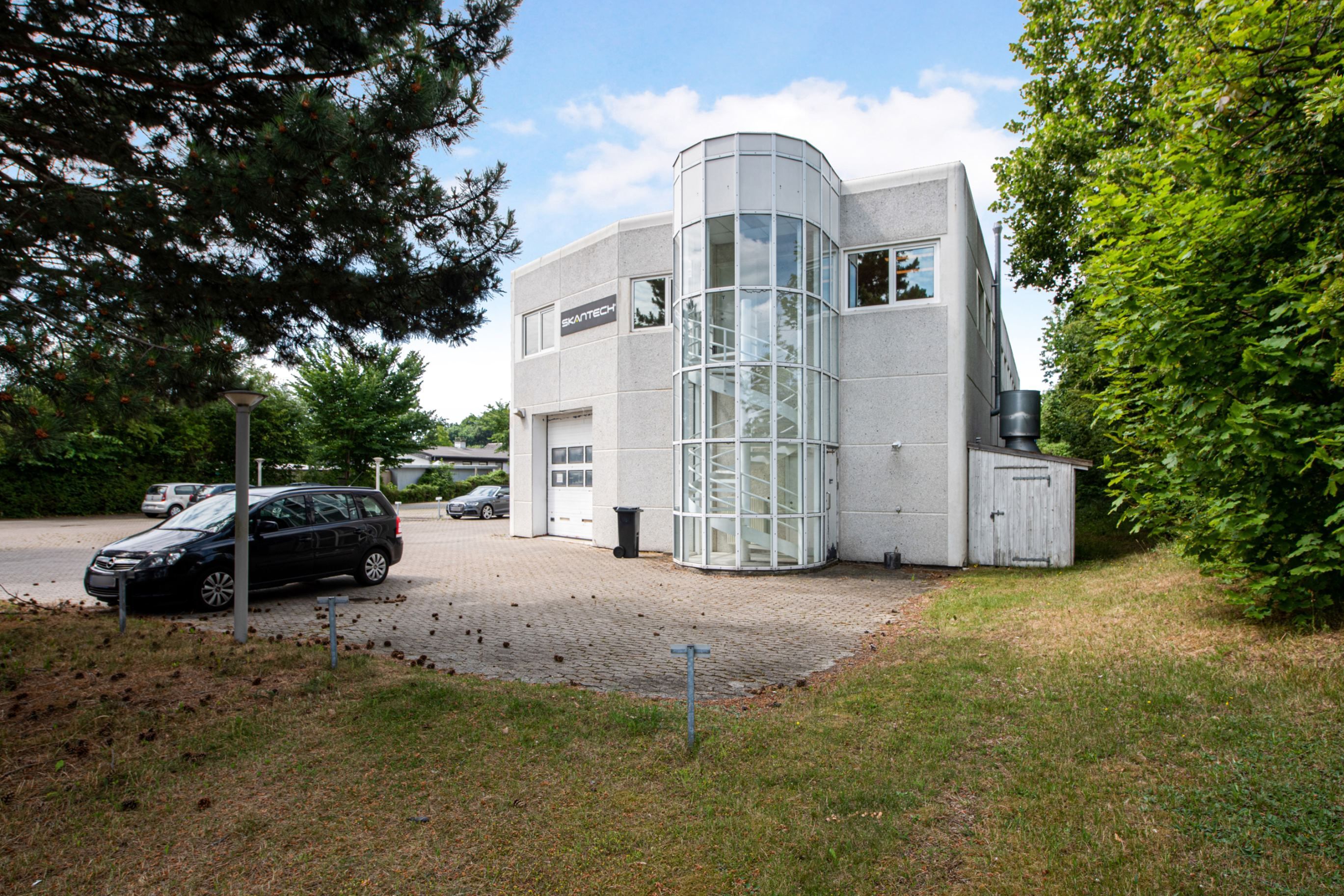 Kontorbyggeri med moderne glasvinduer og højt trappehus. To biler parkeret foran. Skantech logo på facaden. Træer og græs i
