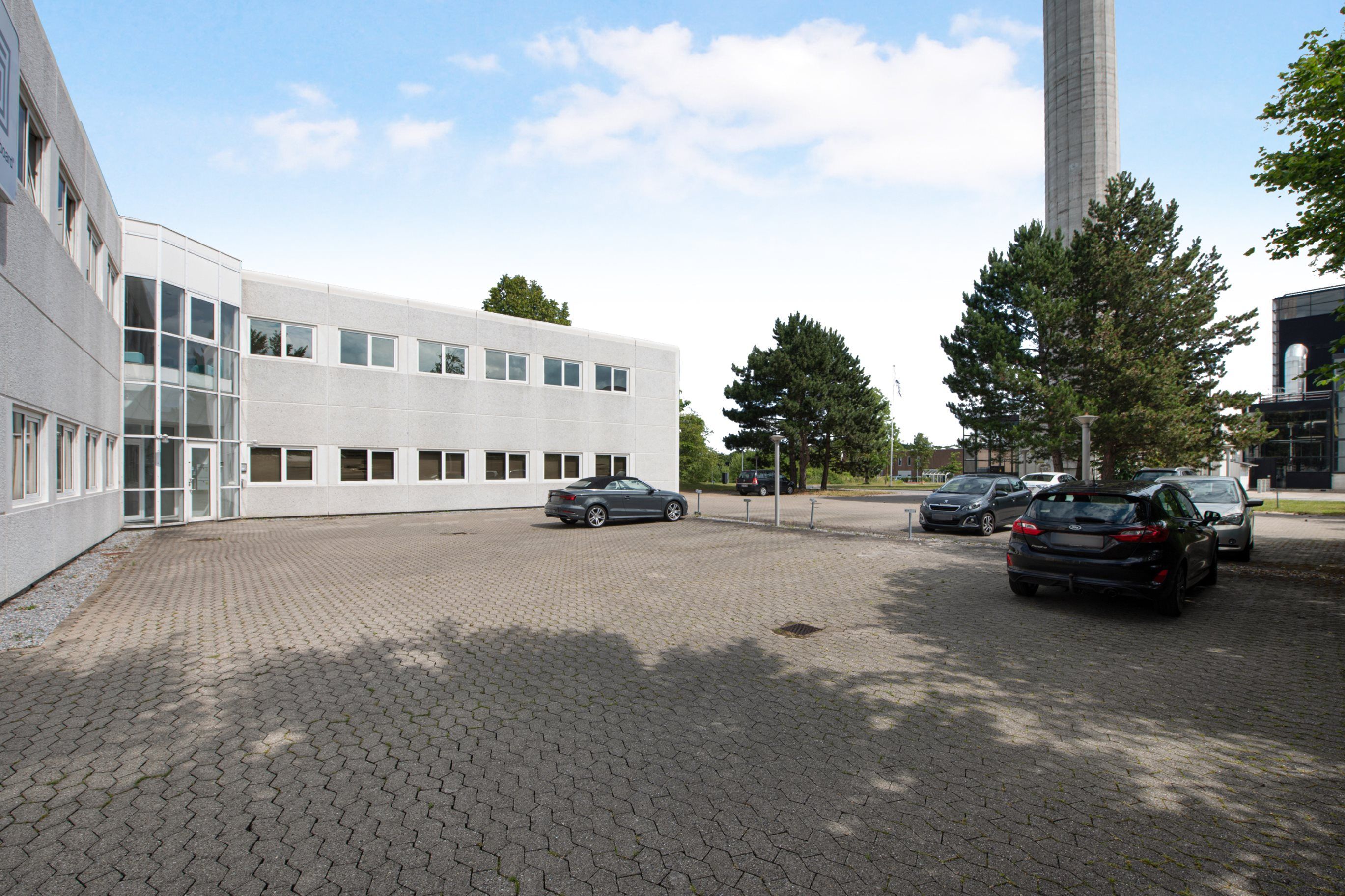 Et kontorlokal med parkeringsplads foran, og der er parkeret biler bag bygningen, og en høj skorsten i baggrunden