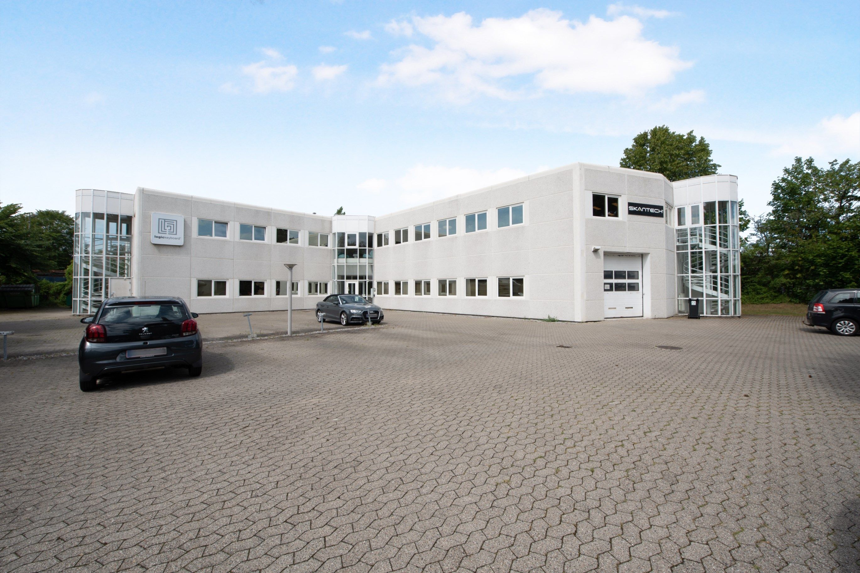 Store hvide bygninger med glasvinduer og parkeringsplads foran, to biler parkeret og et skråt trappeløb