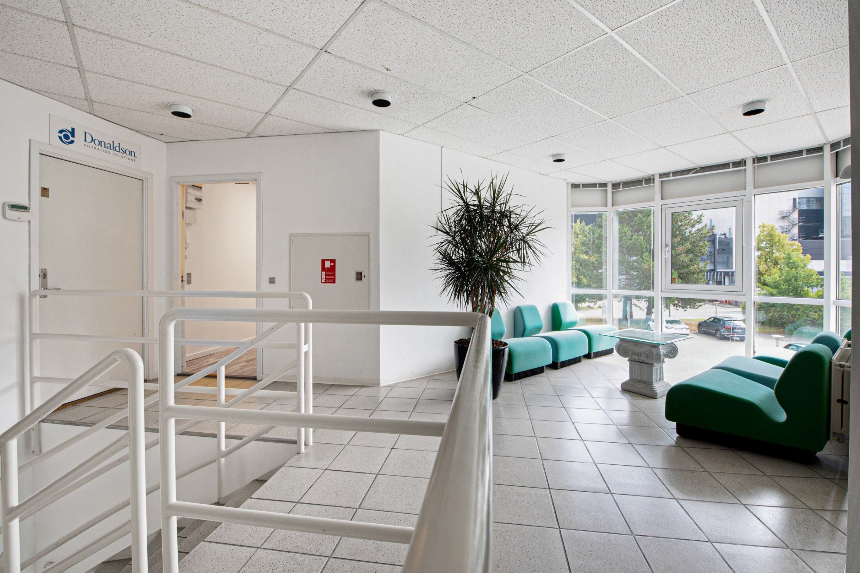 Godt mødested med hvide vægge og gulv, stolpe og håndtag, møbler og plante og vinduer med udsigt til parkeringsplads og biler