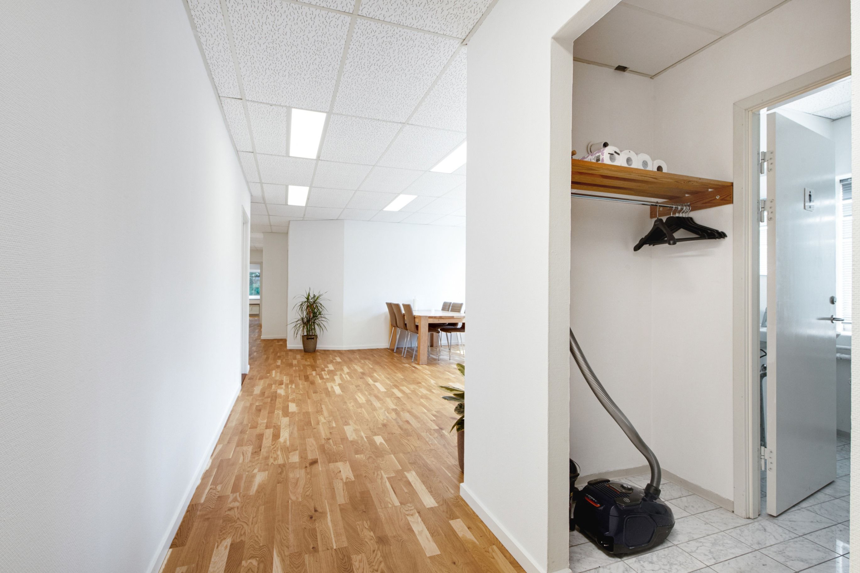 Et kontor med hvide vægge og trægulv samt et lille skab med toiletpapir og hængeklæder