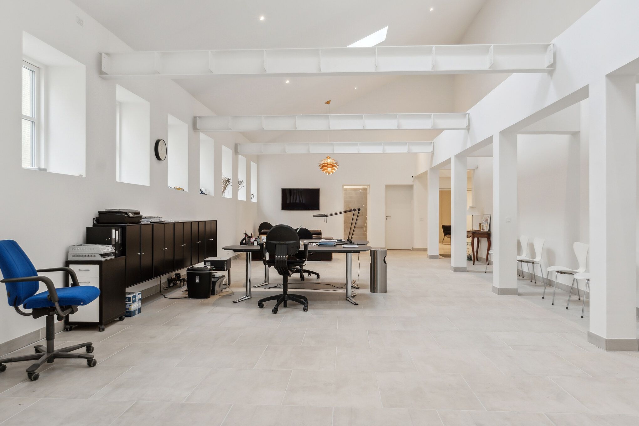 Store, lyse lokaler med højloft, hvide vægge og gulv. Her er to skriveborde med kontorstolar, en skrivemaskine og en