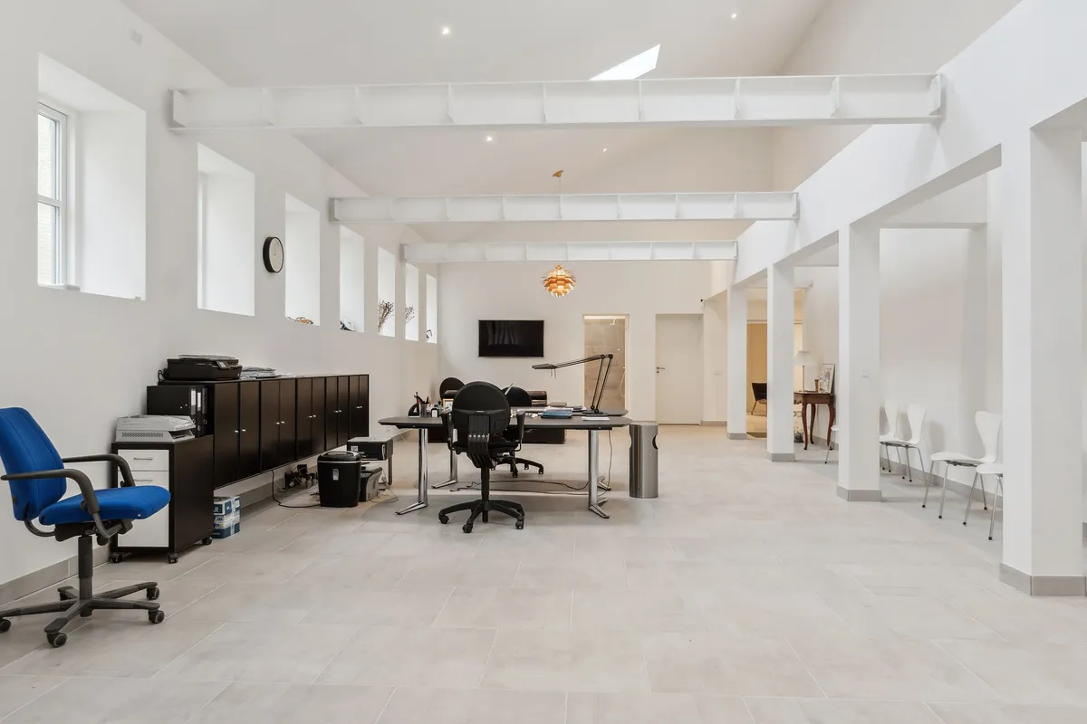 Store, lyse lokaler med højloft, hvide vægge og gulv. Her er to skriveborde med kontorstolar, en skrivemaskine og en