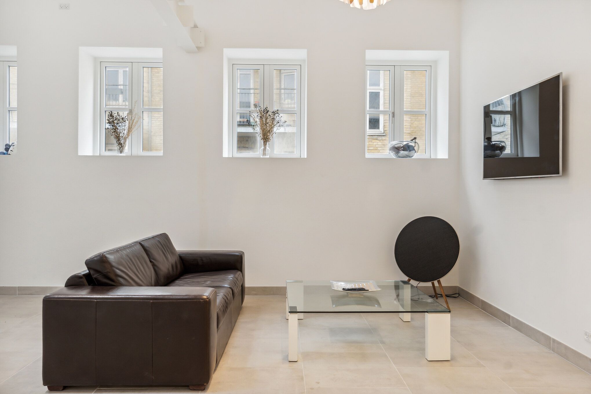 Minimalistisk stue med højloft, hvor tre små ruder viser byudsigt og en fladt skærm i højre sidevæg