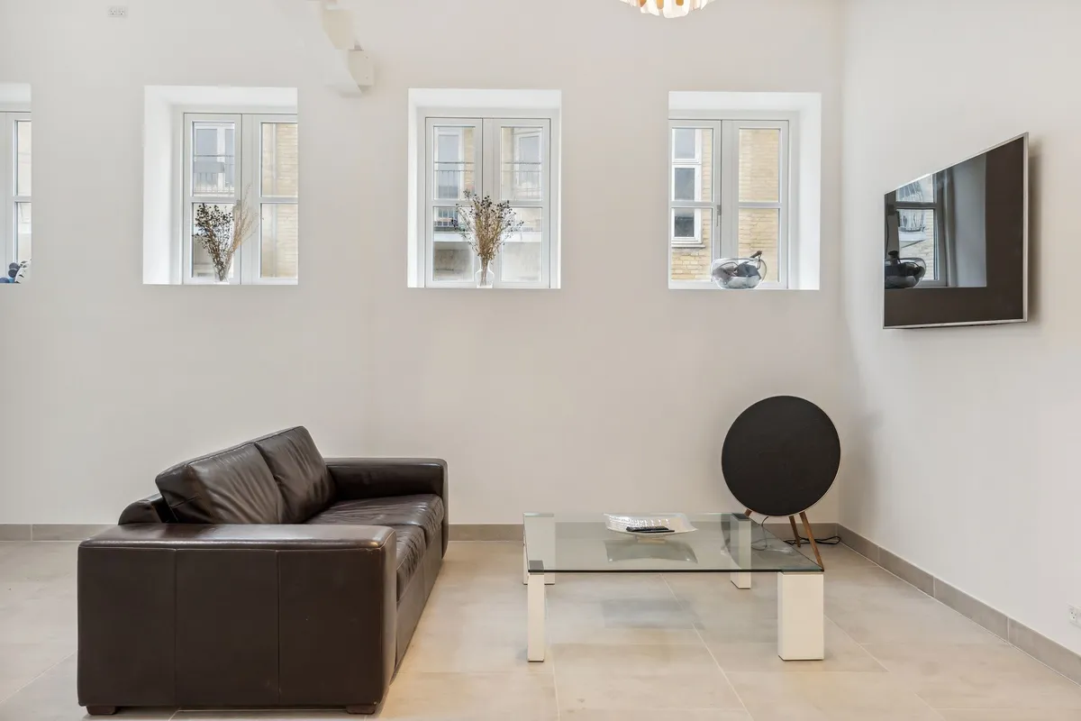 Minimalistisk stue med højloft, hvor tre små ruder viser byudsigt og en fladt skærm i højre sidevæg