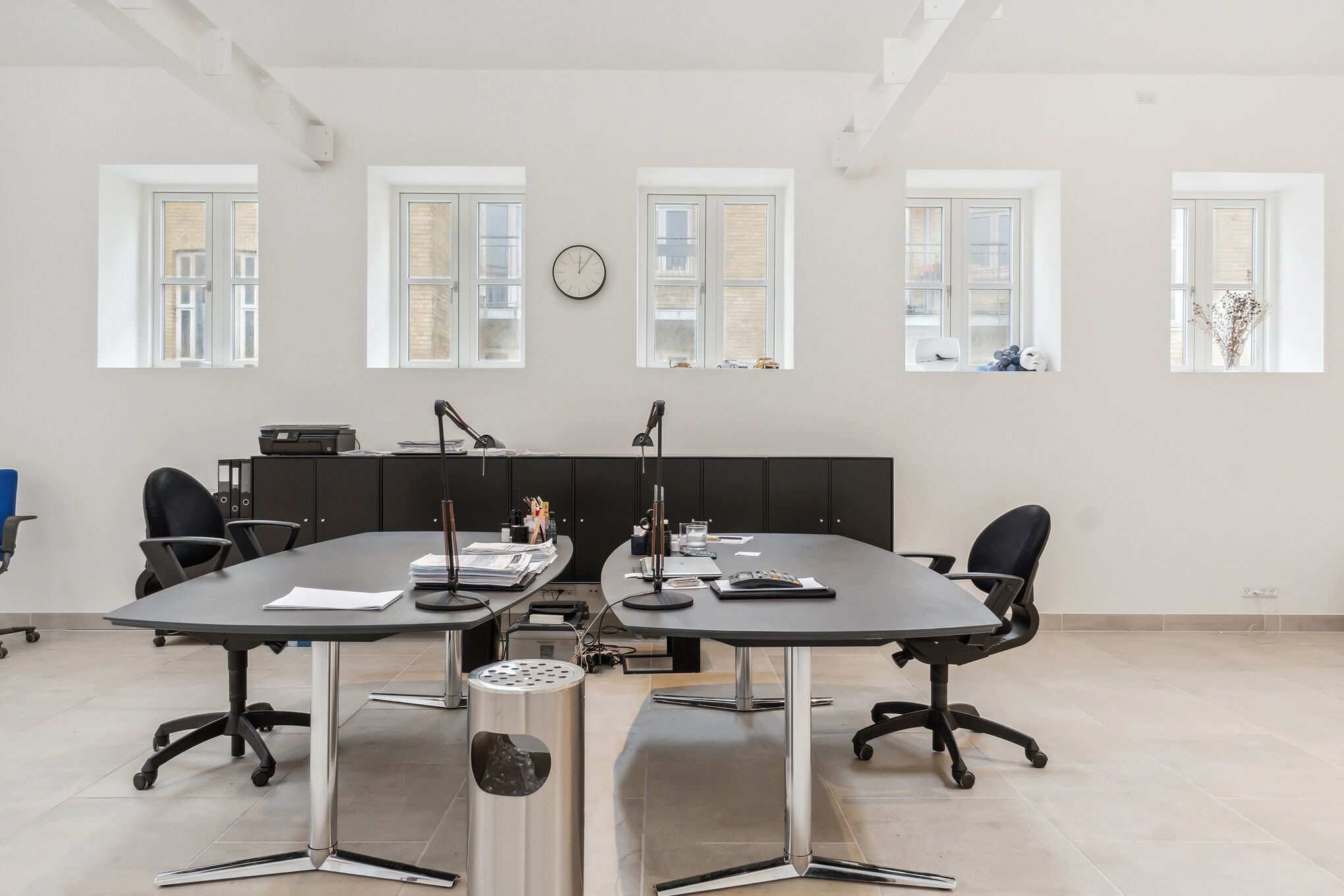 Kontor med to lange borde og mødestoler, højloft og fem store vinduer med kviksølvglas. Bord med aviser og lampe. Sort