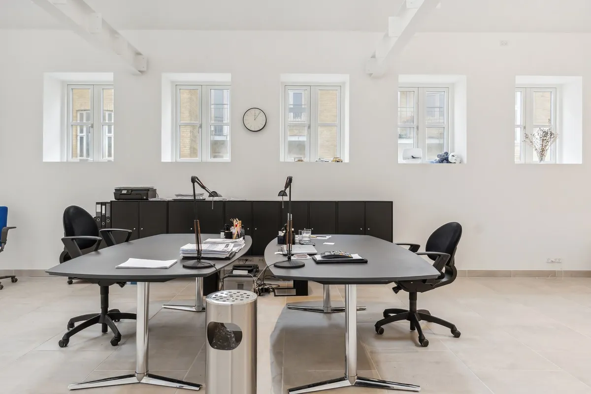 Kontor med to lange borde og mødestoler, højloft og fem store vinduer med kviksølvglas. Bord med aviser og lampe. Sort