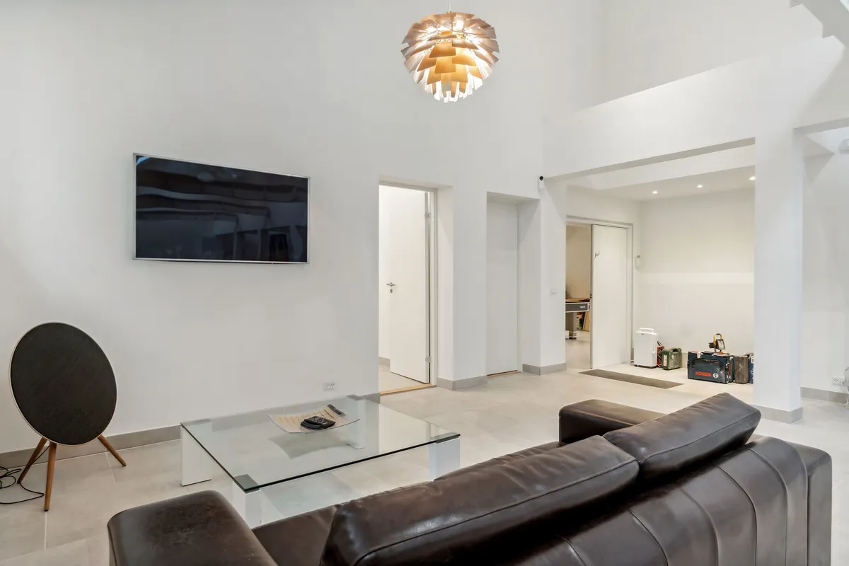 Et rum med hvidt loft og vægge, en stor sort lysekrone, et tv-apparat, en lædersofa og en glasbord med en pude