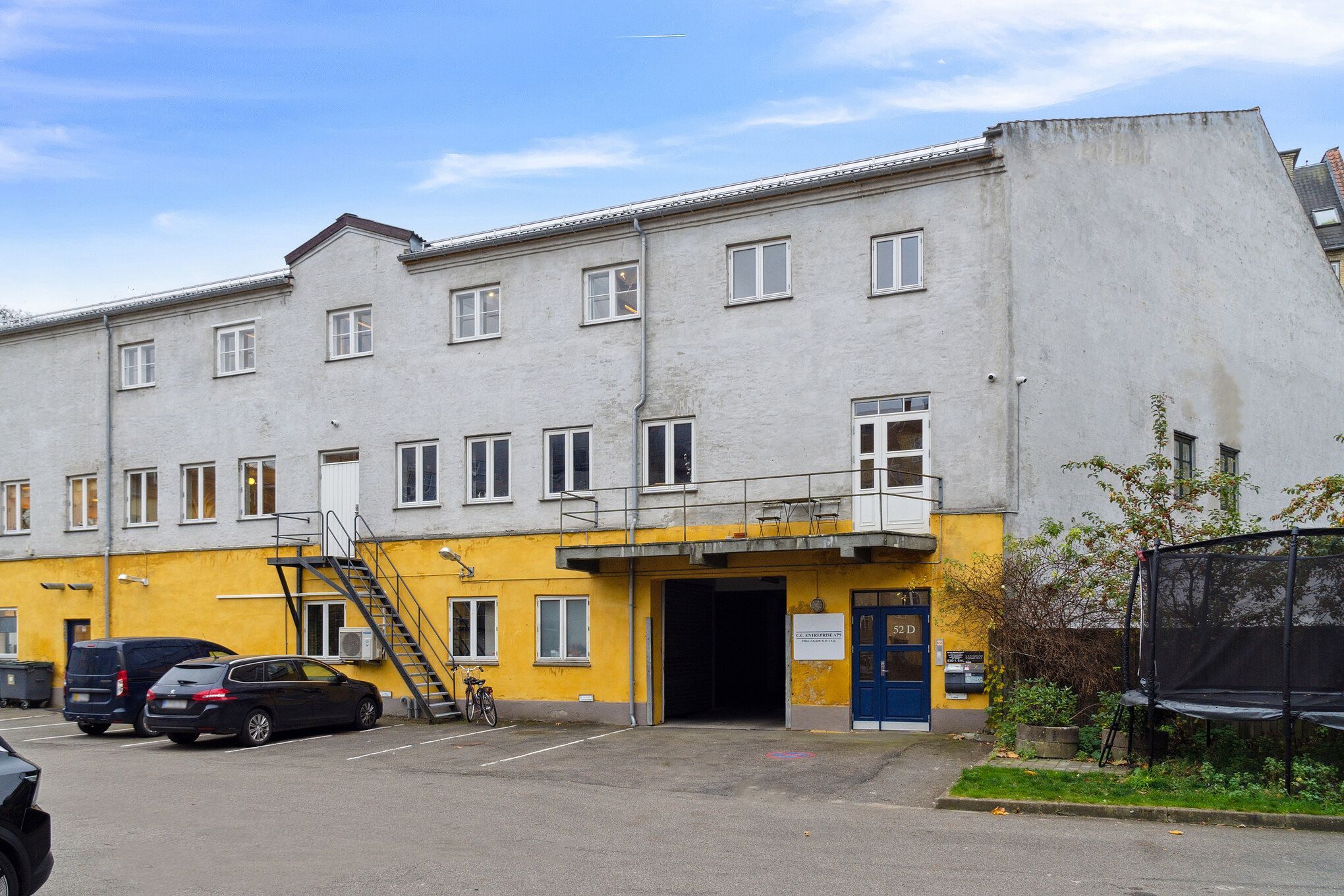 Loft til kip og stor vareelevator til etagen. To biler parkeret i parkeringspladsen. Trampolin og træer ved siden af