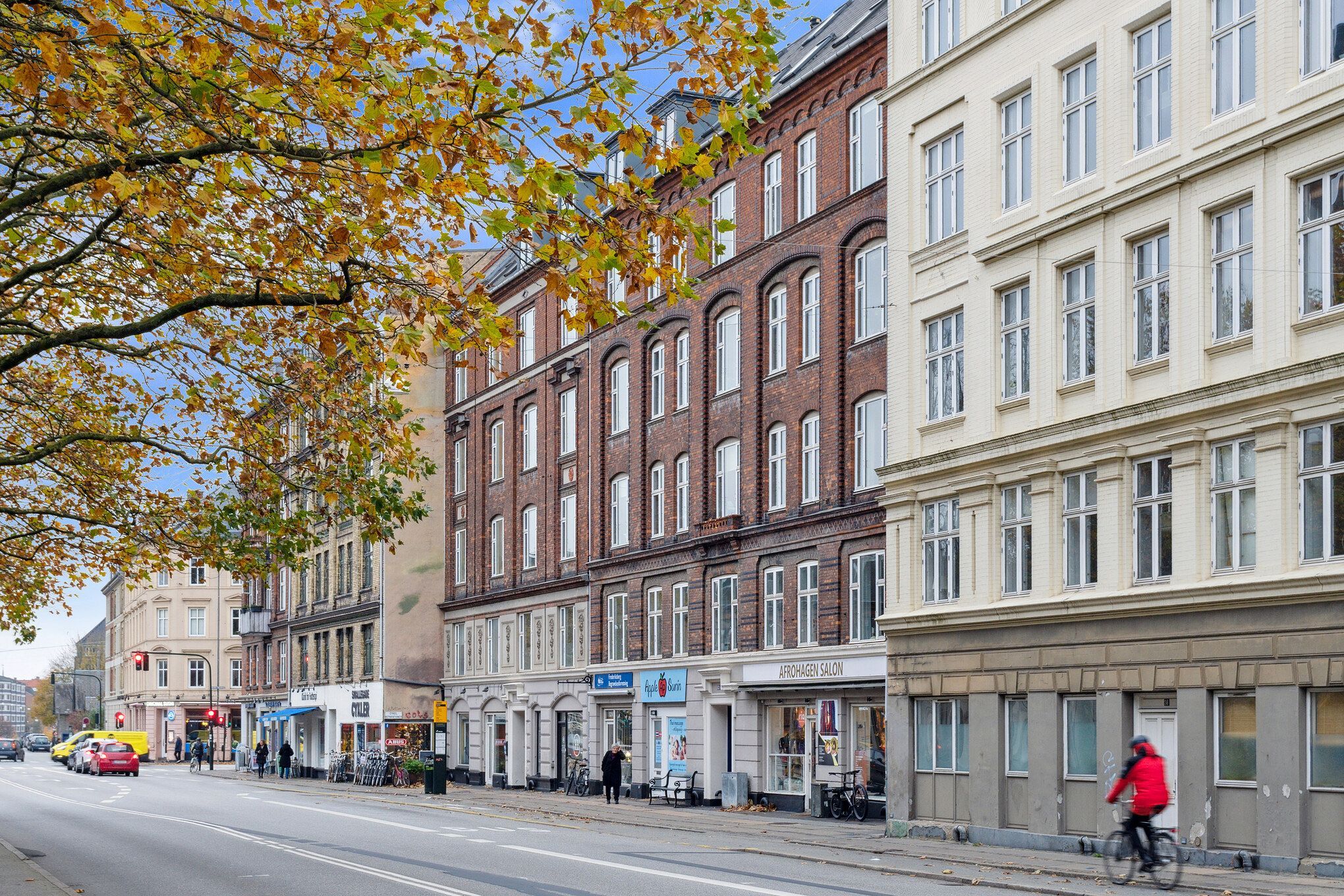En person cykler forbi et stort rødt bygningskompleks med butikker og kontorer i København med røde teglsten og mange vinduer