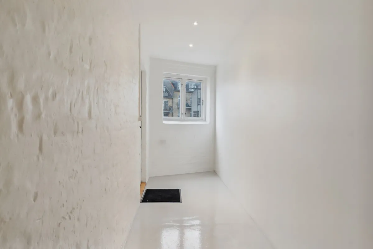 Indvendigt rum med hvide vægge og gulv samt et vindue med udsigt til en bygning og en sort dørmat