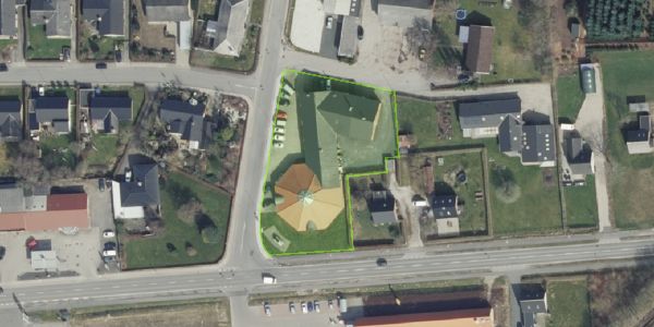 Erhvervsgrund med stort gult pavillon på Svinget 2 i Jelling, omgivet af grønne områder og bebyggelse, med bilparkering og