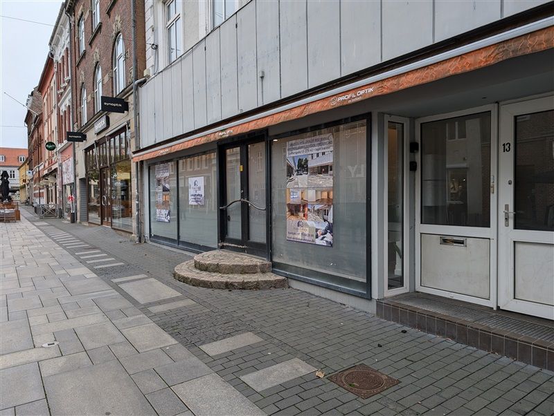 Et butiksvindue med annoncer og skrifttægter i en husrække med stenbrolægget fortov og søjler i baggrunden