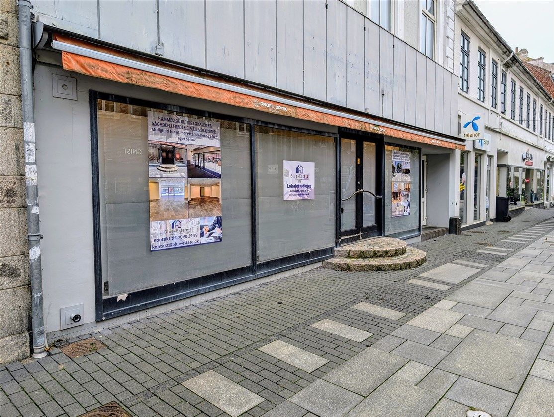 Økonomisk ejendom til leje i Fredericia med vinduer, reklameplakater og en lukket dør i en række med butikker