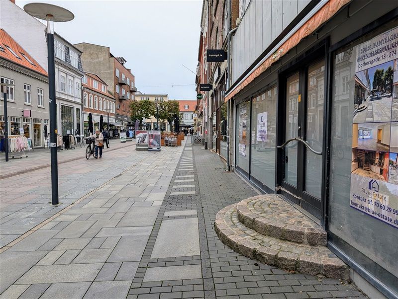 En tom gade med forretningslokaler og cykelstel, søjler og reklamer og mennesker på cykel og gående