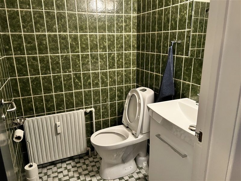 Bad med grønne flisede vægge og toilet med åben låg og hvidt sink. En hængende håndklæde og radiatorer er placeret på væggen