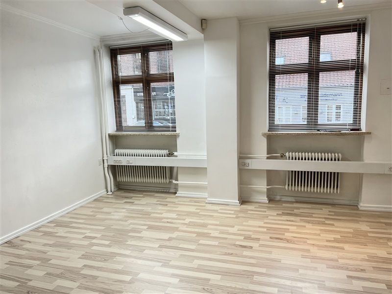 Et rum med to vinduer, hvide vægge og loft og trægulve. To radiatorer med jalousier og lysekroner på loftet.