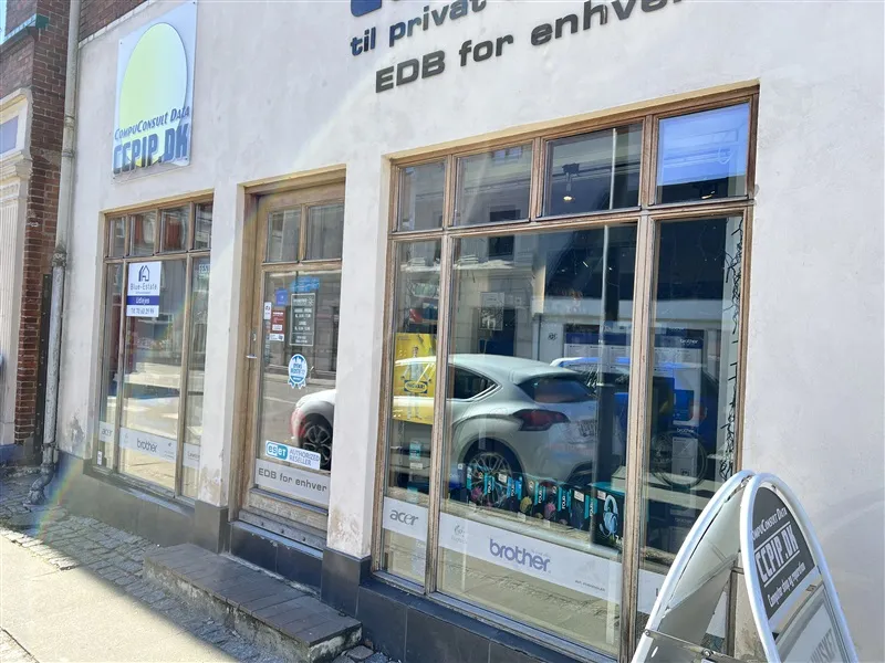 Butiksfront med glasruder og skilte med teksten "EDB for enhver" og "brother" samt en refleksion af bil og vej på glasruderne