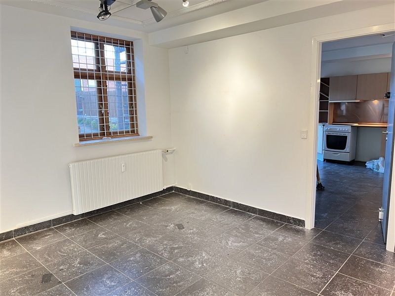 En tom stue med hvide vægge og en radiator, et vindue med et metalgitter og en dør til køkkenet med en hvid komfur