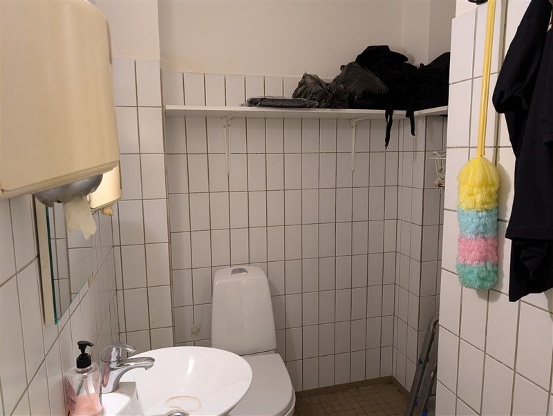 Toiletrum med hvidt toilet, hvidt vask og håndvask med sæbe. Toiletpapirautomat og spejl. Rum med hvide fliser og stige til