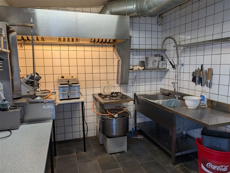 Professionelt køkken med ovn, komfur, blodger, vaskebækken og apparater som kødsliber og ristning.