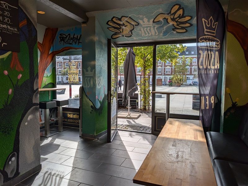 Inde i en restaurant med åbne døre og træbord, der vender mod gaden med planter og bygninger i baggrunden