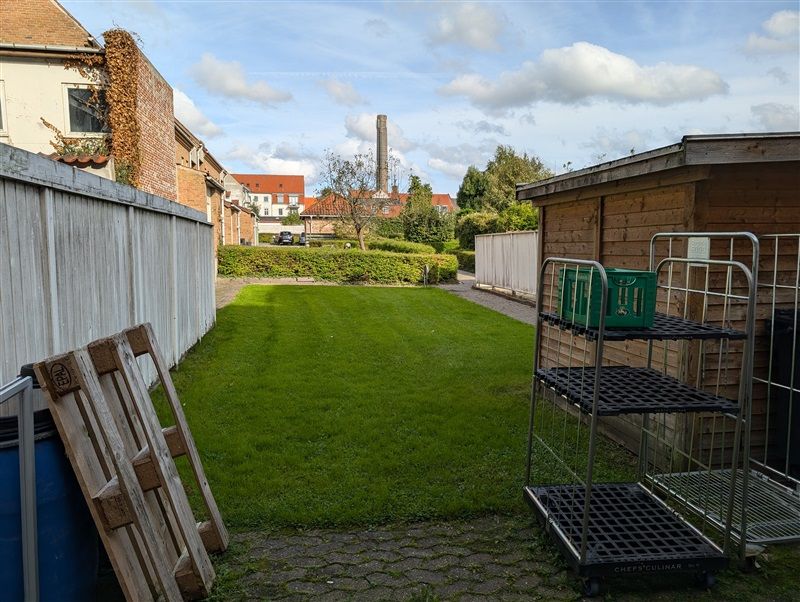 En park med grønne græsflader og buske, stakkelåg og skraldespand, to metalreller med grønne kurve og en gård med røde tag