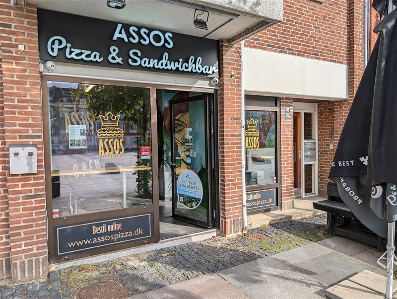 Glassdøre til restauranten med teksten Assos Pizza & Sandwichbar og et maleri af en kvinde og en krone