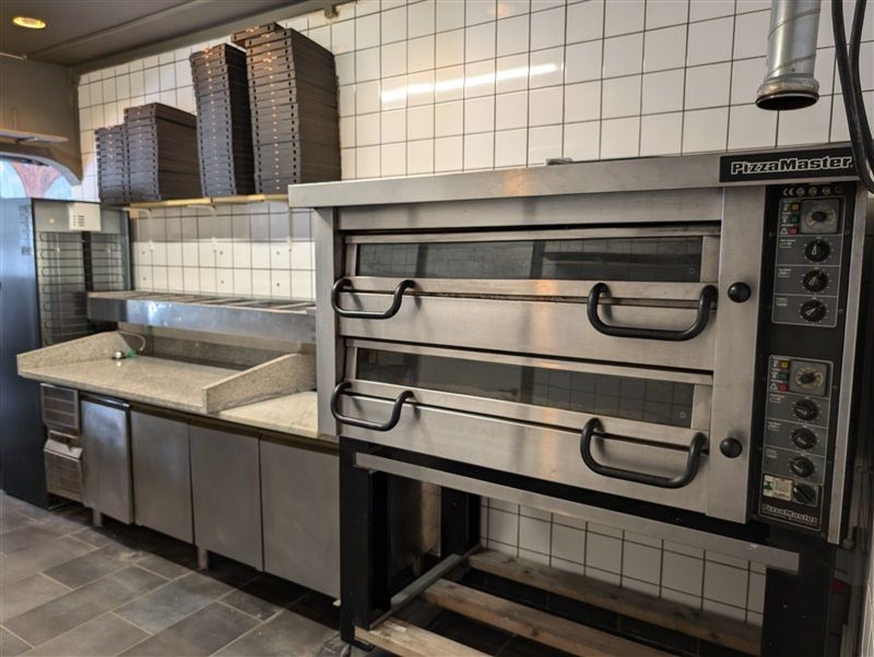 PizzaMaster-ovn med to afdelinger står i et køkken med lysegrå flisede vægge og en udstyret arbejdsbænk med en indbygget