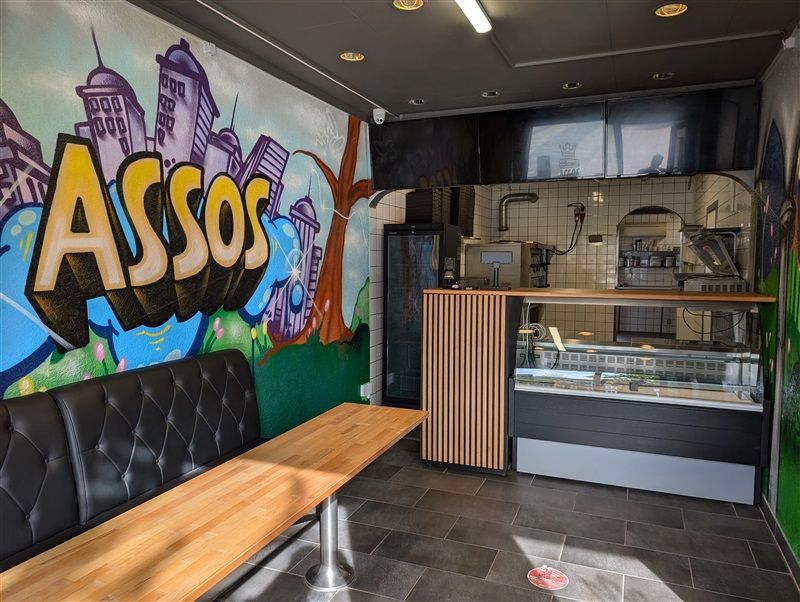 Køkken med servitre, skabe, glasvitrine, skrivebord og sofa i en restaurant med en graffiti-væg med ordet "Assos"