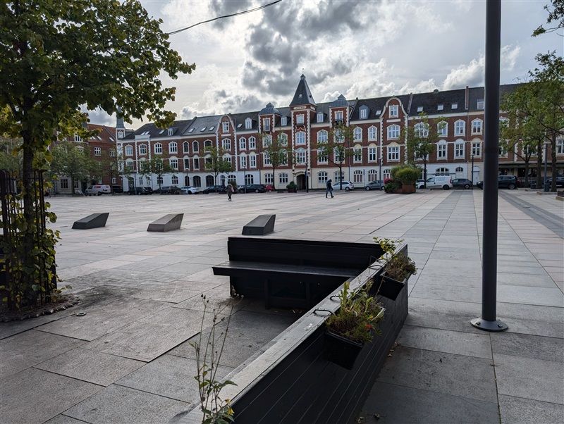 Et tomt parkområde med betonplader, møbler og planter i et byområde med bygninger og biler i baggrunden