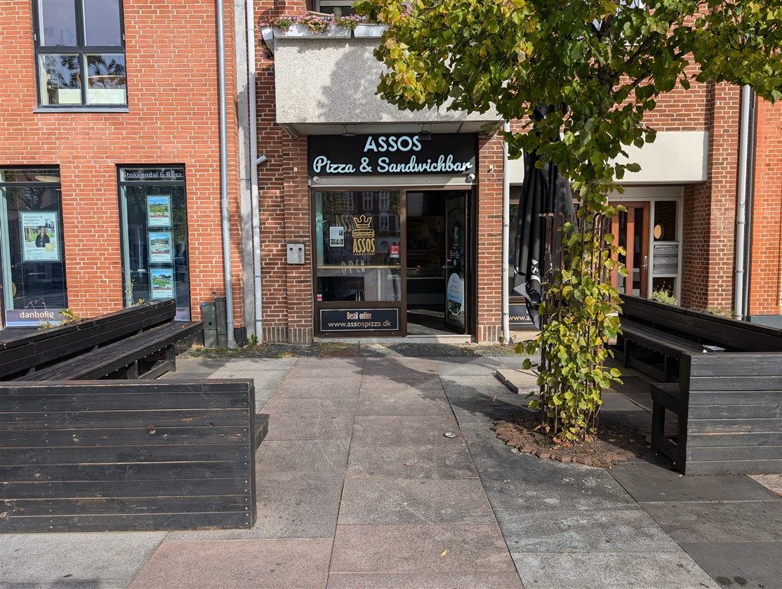 En søjle med en plante i jernbeton, et gulv med flere bænke og en restaurant med navnet Assos Pizza & Sandwichbar