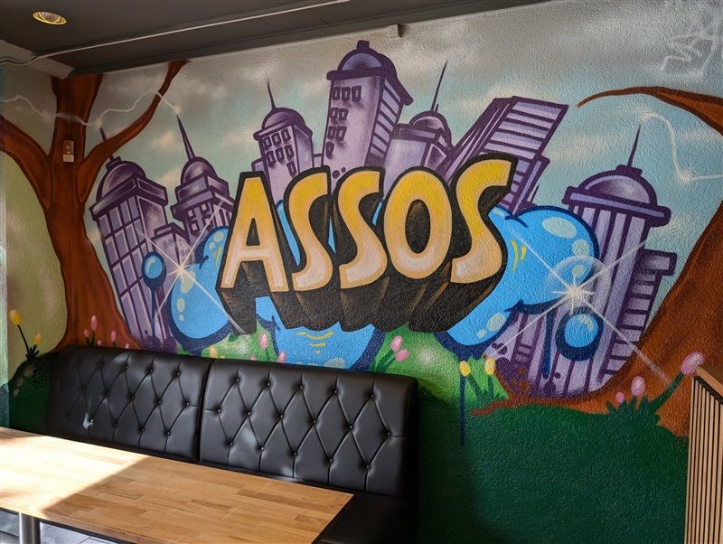 En stol med et bord og en væg med graffiti-kunst, der skriver "ASSOS" med bygninger og grønne planter