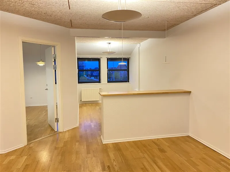 Et skandinavisk interiør med hvide vægge, hårdt trægulv, og moderne belysning samt to vinduer med udsigt til nabobygger og