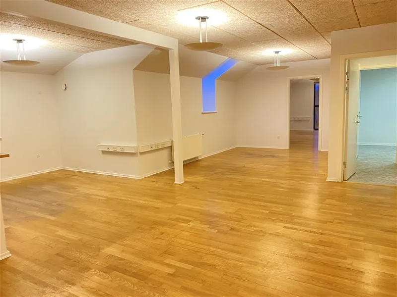 Et tomt kontor med massiv trægulv, højt loft, åbne døre og tre lysekroner hængende fra loftet