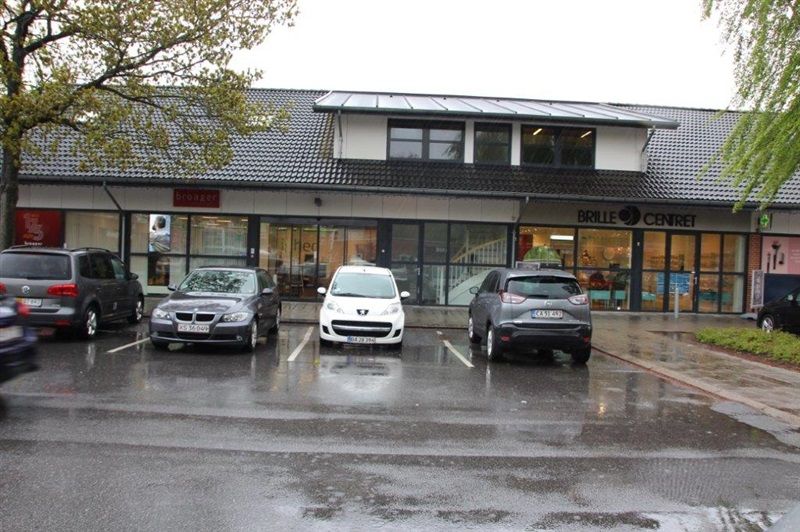 Forretningspark med forretninger, Brille Centret og Broager. Fire biler parkeret foran bygningen i regnvejr