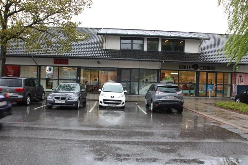 Forretningspark med forretninger, Brille Centret og Broager. Fire biler parkeret foran bygningen i regnvejr