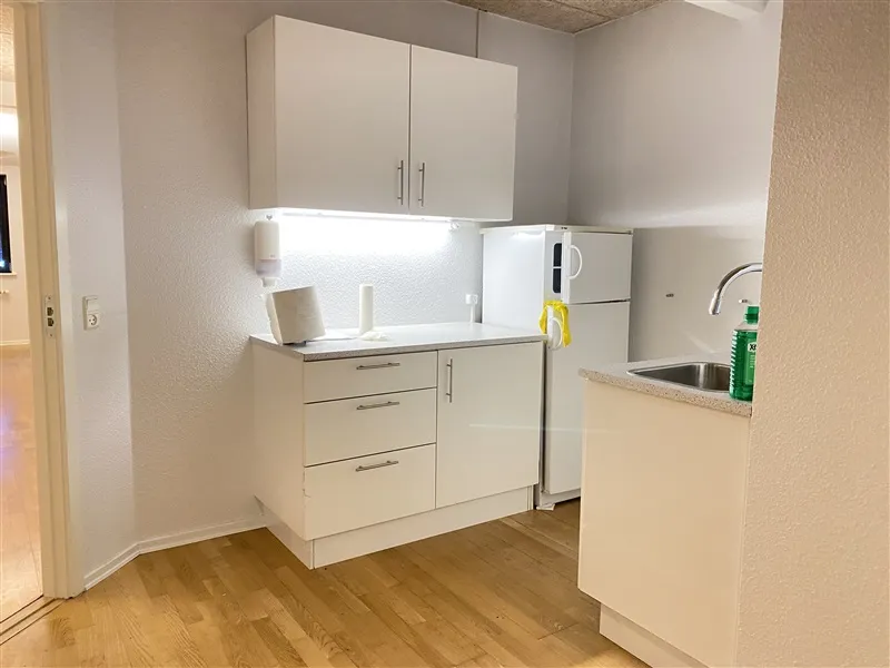 Moderne køkken med hvide vægge, køkkenskabe og hætte over komfuret. Et hvidt køleskab står mod væggen, og der er en vask med