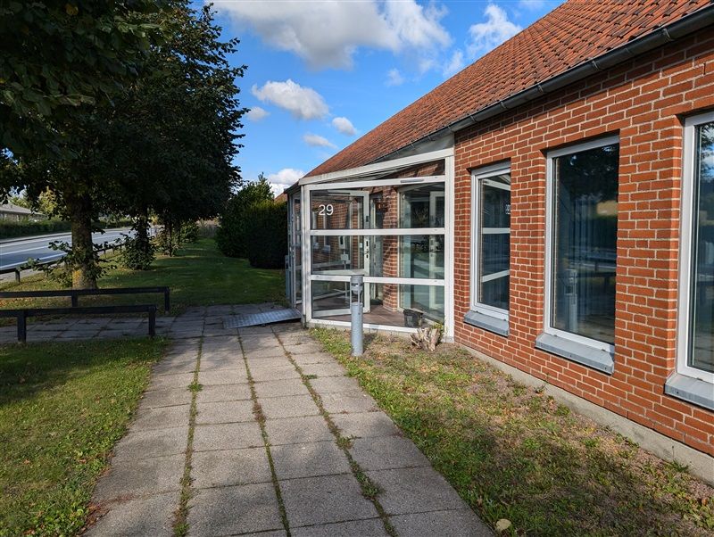 En erhvervsejendom med nummer 29 og en åben dør og vinduer i røde mursten langs en grønsnethældt sidevej med træer