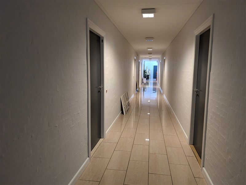 En lang korridor med hvide vægge og lysgrå gulvfliser, to døre og lyskroner i loftet