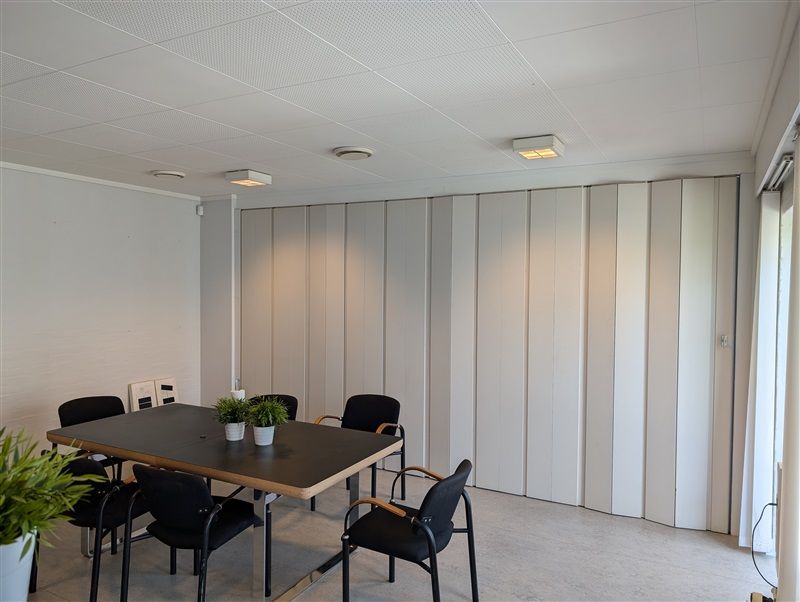 Ny mødelokale med højt til loftet, lyse hvide vægge og en stor foldeluge. Bord med to potter og en lænestol og stol til hver