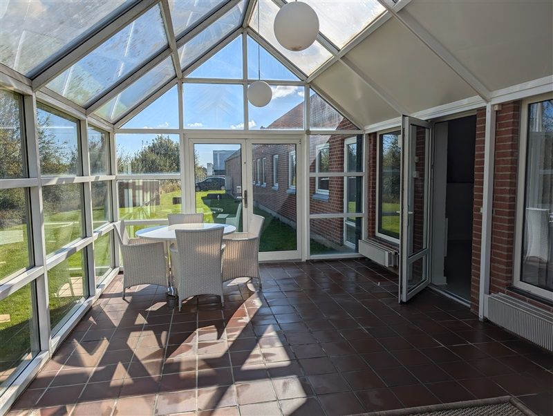 Terrasse med glasvinduer, hvide stole og et rundt bord placeret midt på terrassen. På loftet hænger to hvide kuglelys.